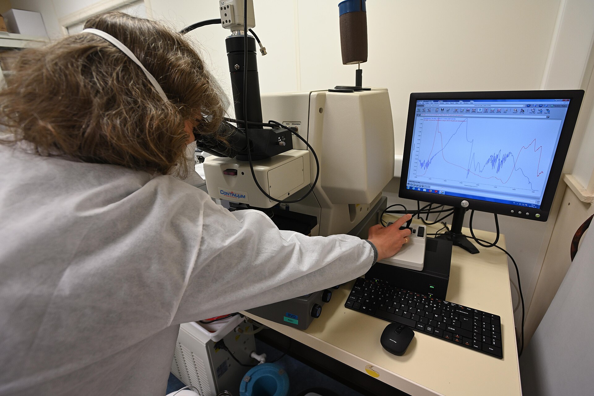 Scientist in a lab coat reviewing overlaid infrared spectra in spectroscopy analysis software on a monitor next to a microscope-coupled spectrometer